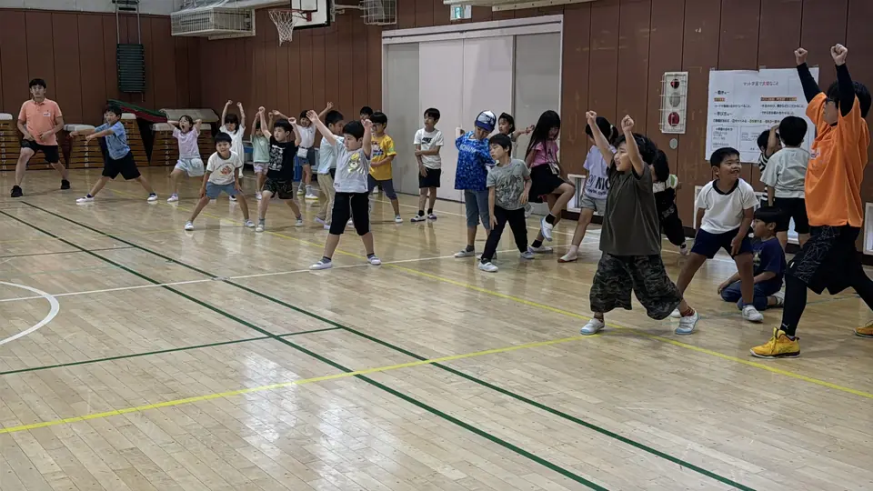 小学校の体育館で大人数の児童が一斉に身体を動かす様子