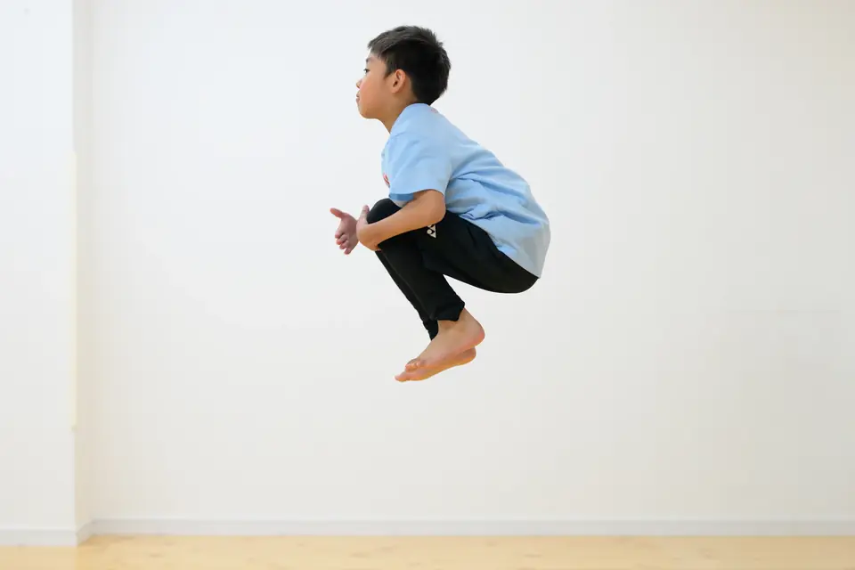 白い壁を背景に、両脚を抱えて高く跳ぶ子ども